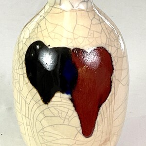 May include: A cream-colored ceramic vase with a crackle glaze. The vase features a design of three overlapping shapes in black, blue, and red. The vase has a rounded body and a narrow neck.