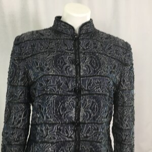 K&ouml;nnte beinhalten: Dunkelblaue Jacke mit hohem Kragen und durchgehendem Rei&szlig;verschluss. Die Jacke hat ein kompliziertes, strukturiertes Muster mit schwarzem Besatz. Das Design umfasst florale und geometrische Formen. Die Jacke wird auf einer wei&szlig;en Schaufenquin pr&auml;sentiert.