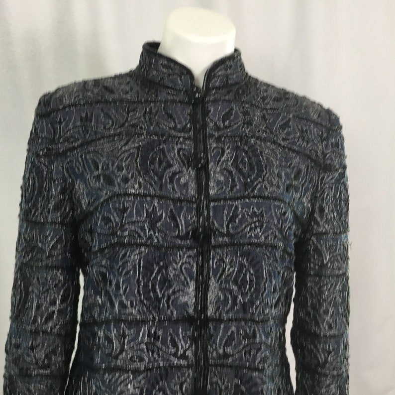 K&ouml;nnte beinhalten: Dunkelblaue Jacke mit hohem Kragen und durchgehendem Rei&szlig;verschluss. Die Jacke hat ein kompliziertes, strukturiertes Muster mit schwarzem Besatz. Das Design umfasst florale und geometrische Formen. Die Jacke wird auf einer wei&szlig;en Schaufenquin pr&auml;sentiert.