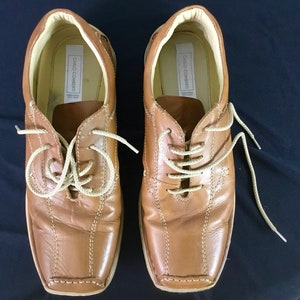 May include: A pair of brown leather lace-up shoes with a square toe. The shoes have a cream-colored lace and the brand name "Carlo Contenti" is visible on the inside of the shoes.