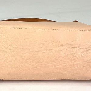 May include: A light pink leather handbag with a brown handle. The bottom of the bag has four gold-colored metal feet. The leather has a textured appearance.