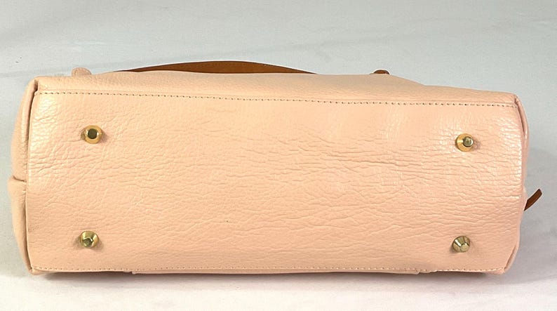 May include: A light pink leather handbag with a brown handle. The bottom of the bag has four gold-colored metal feet. The leather has a textured appearance.