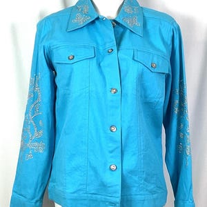 May include: A turquoise denim jacket with silver floral embellishments on the collar and sleeves. The jacket has two chest pockets and silver button closures. The long-sleeved jacket is displayed on a mannequin.