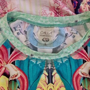 Op de afbeelding: Een groen-wit gestreept shirt met een label met de tekst "CALLA by Carrie Underwood" en een maatlabel "L". Het shirt heeft een kleurrijke bloemenprint met rode, gele en blauwe bloemen.