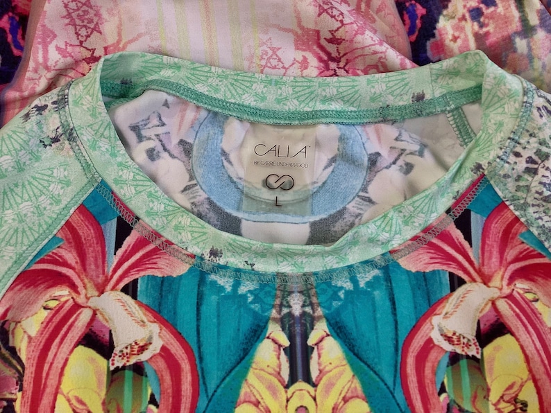 Op de afbeelding: Een groen-wit gestreept shirt met een label met de tekst "CALLA by Carrie Underwood" en een maatlabel "L". Het shirt heeft een kleurrijke bloemenprint met rode, gele en blauwe bloemen.