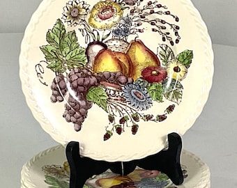 May include: A stack of cream-colored ceramic plates with a fruit and floral design. The plates feature a detailed illustration of grapes, pears, and flowers. Each plate has a decorative, textured rim.
