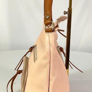 May include: A light pink leather handbag with a brown leather handle. The bag has a side pocket with a zipper and brown leather accents. The bag is displayed on a gold-colored stand.