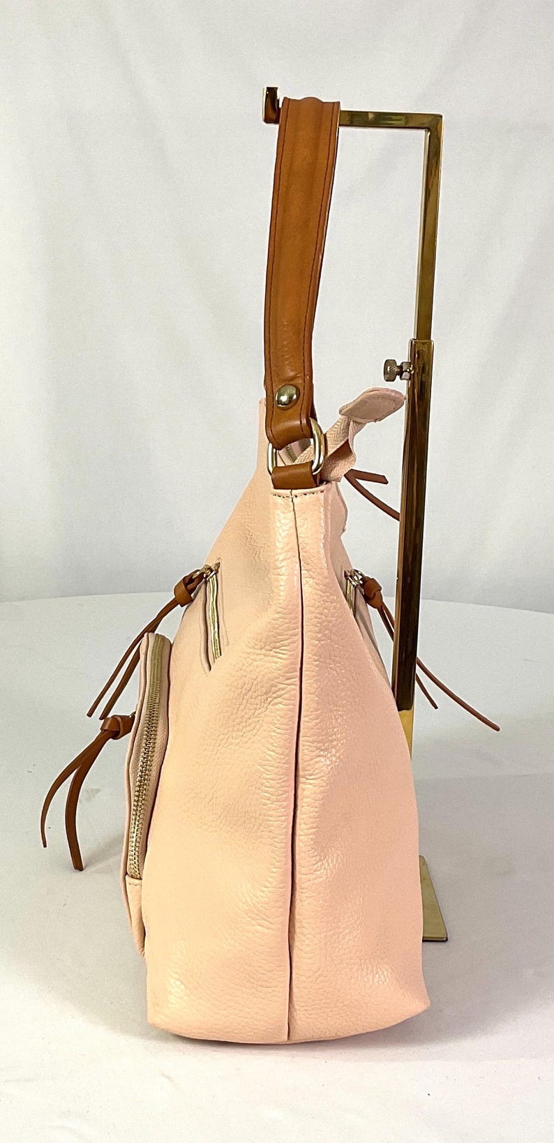May include: A light pink leather handbag with a brown leather handle. The bag has a side pocket with a zipper and brown leather accents. The bag is displayed on a gold-colored stand.