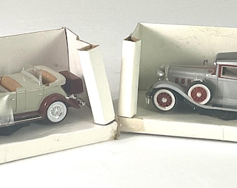 May include: Two vintage model cars in their original boxes. One is a convertible with a light green body, brown accents, and white-walled tires. The other is a gray sedan with red accents and white-walled tires. Both are displayed in open cardboard boxes.