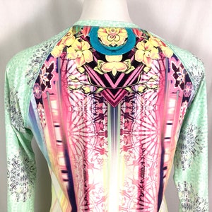 Op de afbeelding: Een shirt met lange mouwen met een kleurrijke, abstracte print. Het shirt heeft een groene basiskleur met een veelkleurig ontwerp met geometrische vormen, bloemen en een regenboogachtig patroon.