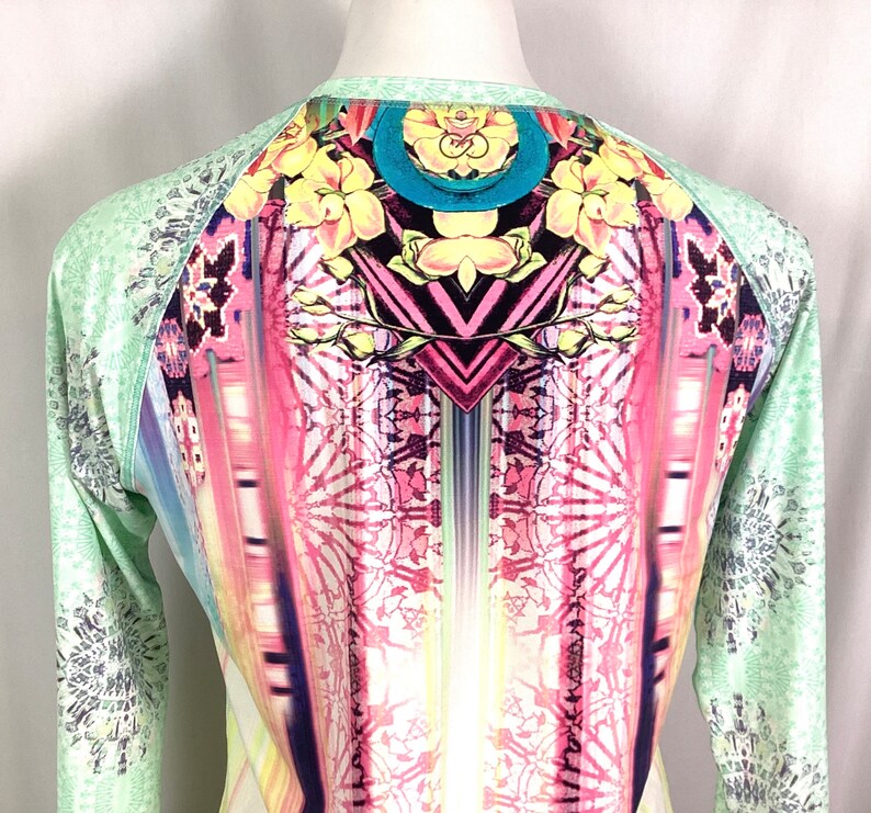 Op de afbeelding: Een shirt met lange mouwen met een kleurrijke, abstracte print. Het shirt heeft een groene basiskleur met een veelkleurig ontwerp met geometrische vormen, bloemen en een regenboogachtig patroon.