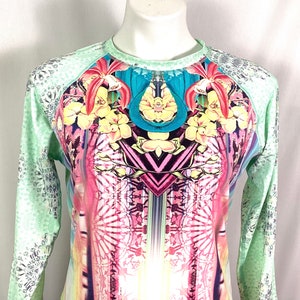 Op de afbeelding: Een shirt met lange mouwen met een kleurrijke, abstracte print met bloemen, geometrische vormen en een turquoise ontwerp. Het shirt heeft een lichtgroene achtergrond met een wit en grijs patroon op de mouwen.