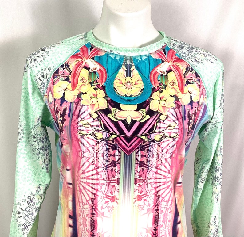 Op de afbeelding: Een shirt met lange mouwen met een kleurrijke, abstracte print met bloemen, geometrische vormen en een turquoise ontwerp. Het shirt heeft een lichtgroene achtergrond met een wit en grijs patroon op de mouwen.