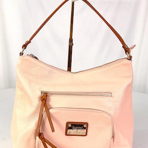 May include: A light pink leather shoulder bag with brown leather straps. The bag has a front pocket with a zipper and a decorative leather tassel. A metal plaque with the text "Valentina Italia" is on the front.