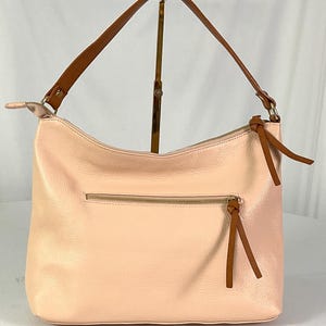 May include: A light pink leather shoulder bag with a brown strap and zipper. The bag has a textured surface and a brown tassel on the zipper. The bag is displayed on a gold stand against a white background.