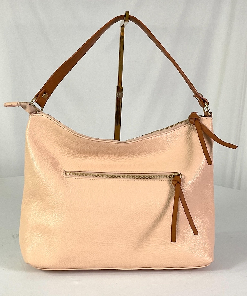 May include: A light pink leather shoulder bag with a brown strap and zipper. The bag has a textured surface and a brown tassel on the zipper. The bag is displayed on a gold stand against a white background.