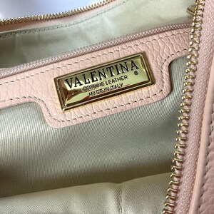May include: Close-up of a light pink leather handbag interior. A gold-colored rectangular plaque reads "VALENTINA GENUINE LEATHER MADE IN ITALY". The bag has a gold zipper and a light-colored fabric lining.