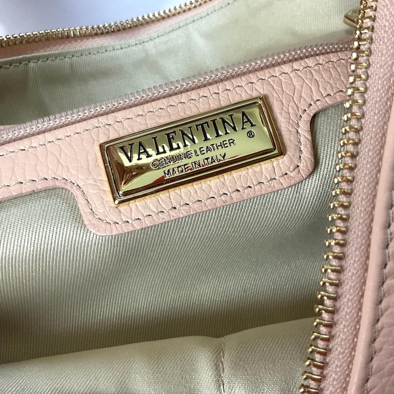 May include: Close-up of a light pink leather handbag interior. A gold-colored rectangular plaque reads "VALENTINA GENUINE LEATHER MADE IN ITALY". The bag has a gold zipper and a light-colored fabric lining.