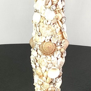 Puede incluir: Un portavelas blanco hecho completamente de conchas marinas. El portavelas es alto y cilíndrico con una base cuadrada. Las conchas marinas son de varios tamaños y formas, y están dispuestas en un patrón aleatorio.