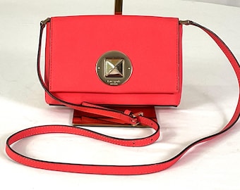May include: A coral-colored crossbody bag with a gold-tone clasp. The bag features a rectangular shape with a flap closure and a long, thin strap. The clasp has a square design with a pyramid-shaped center.