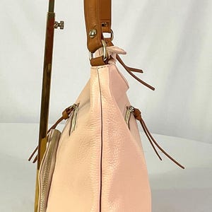May include: A light pink leather handbag with a brown leather strap. The bag has a soft, textured appearance and features multiple zippered compartments. The bag is displayed on a gold-colored stand.