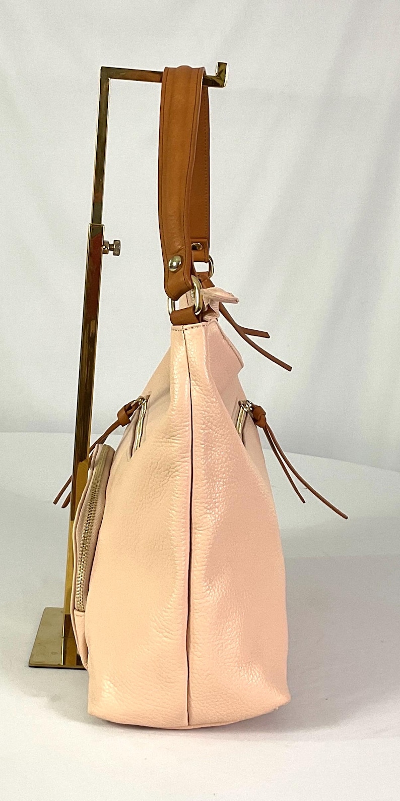May include: A light pink leather handbag with a brown leather strap. The bag has a soft, textured appearance and features multiple zippered compartments. The bag is displayed on a gold-colored stand.