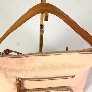 May include: A light pink leather handbag with a brown leather handle. The bag has two zippered pockets on the front and a gold zipper closure. The handle is attached to the bag with gold hardware.