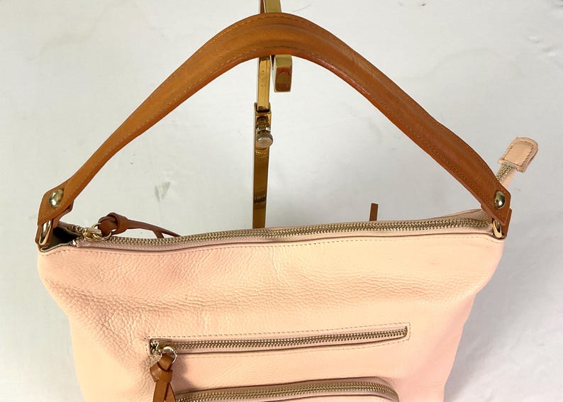 May include: A light pink leather handbag with a brown leather handle. The bag has two zippered pockets on the front and a gold zipper closure. The handle is attached to the bag with gold hardware.