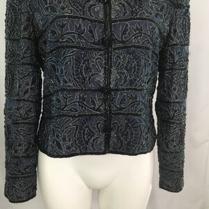 K&ouml;nnte beinhalten: Dunkelblaue Jacke mit aufwendiger schwarzer Perlenarbeit und floralen Mustern. Die Jacke hat eine Knopfleiste und lange &Auml;rmel. Das Design zeigt horizontale Streifen und detaillierte Blumenmotive, die einen strukturierten und eleganten Look erzeugen.