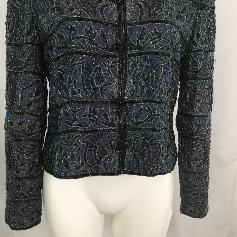 K&ouml;nnte beinhalten: Dunkelblaue Jacke mit aufwendiger schwarzer Perlenarbeit und floralen Mustern. Die Jacke hat eine Knopfleiste und lange &Auml;rmel. Das Design zeigt horizontale Streifen und detaillierte Blumenmotive, die einen strukturierten und eleganten Look erzeugen.