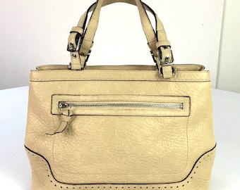 May include: A cream-colored leather handbag with two top handles and a zippered pocket on the front. The bag has a dotted trim around the bottom edge.