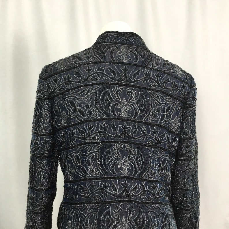 K&ouml;nnte beinhalten: Eine marineblaue Jacke mit aufwendigen Blumen- und Blattmustern, verziert mit silbernen und schwarzen Perlen. Die Jacke hat einen Stehkragen und lange &Auml;rmel. Die R&uuml;ckseite der Jacke zeigt die detaillierte Perlenarbeit und das Design.