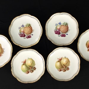 May include: Set of six white porcelain dessert plates with gold trim. Each plate features a different fruit design, including pears, apples, grapes, and pineapple.