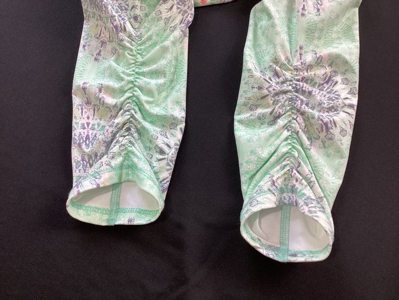 Op de afbeelding: Een paar groene en witte leggings met een patroon en een geplooide detail aan de onderkant van elk been.