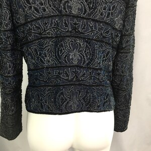 K&ouml;nnte beinhalten: Eine marineblaue Jacke mit aufwendiger schwarzer Perlenarbeit. Die Jacke hat horizontale B&auml;nder mit detaillierten floralen und geometrischen Mustern. Die lang&auml;rmlige Jacke wird auf einer wei&szlig;en Schaufensterpuppe gezeigt, die den R&uuml;cken zeigt.