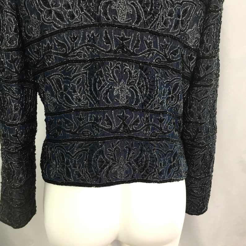 K&ouml;nnte beinhalten: Eine marineblaue Jacke mit aufwendiger schwarzer Perlenarbeit. Die Jacke hat horizontale B&auml;nder mit detaillierten floralen und geometrischen Mustern. Die lang&auml;rmlige Jacke wird auf einer wei&szlig;en Schaufensterpuppe gezeigt, die den R&uuml;cken zeigt.