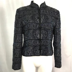 K&ouml;nnte beinhalten: Dunkelblaue Jacke mit Stehkragen und Rei&szlig;verschluss. Die Jacke ist mit aufwendigen floralen Perlenarbeiten verziert und hat lange &Auml;rmel. Das Design weist horizontale Streifen und vertikale Blumenmuster auf.