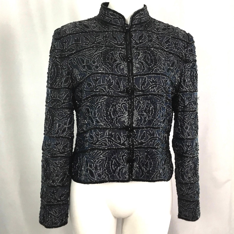 K&ouml;nnte beinhalten: Dunkelblaue Jacke mit Stehkragen und Rei&szlig;verschluss. Die Jacke ist mit aufwendigen floralen Perlenarbeiten verziert und hat lange &Auml;rmel. Das Design weist horizontale Streifen und vertikale Blumenmuster auf.