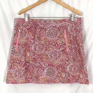 Op de afbeelding: Een roze en rode skort met paisley-print en twee roze zakken met ritssluiting. De skort hangt aan een houten kleerhanger tegen een witte achtergrond. Het patroon bevat bloem- en bladachtige ontwerpen.