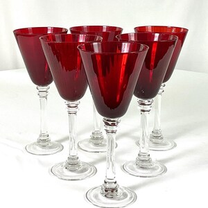 May include: Set of six ruby red wine glasses. Each glass features a clear stem and base, with a deep red bowl. The glasses are arranged in a semi-circular pattern, showcasing their elegant design. Perfect for serving wine at a dinner party.