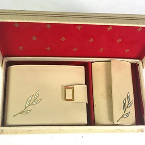 May include: An open vintage gift box with a red interior and gold accents. Inside, a cream-colored wallet with a gold clasp and leaf design, a cylindrical item, and a rectangular item with a leaf design are visible.