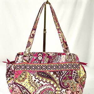 May include: A quilted tote bag with a paisley pattern in shades of pink, purple, and yellow. The bag has two handles and a zippered top. The bag is displayed on a gold-colored stand.