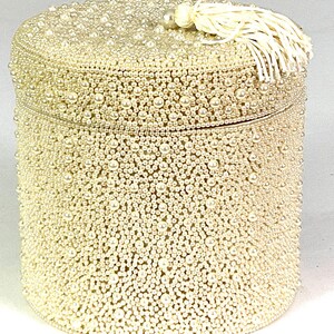 May include: A cylindrical, cream-colored box covered in small, round, faux pearls. The lid is adorned with a pearl and a tassel. The box is a decorative item, possibly for jewelry or small keepsakes.