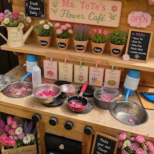 以下が含まれることがあります： 「Ms. TeTe's Flower Café」の看板がある木製のフラワーカフェ。カフェには鉢植えの花、メニュー、そして「Rose Petal Pie」のような特別料理の看板があります。「Pick Your Flowers」と書かれた水やり缶。