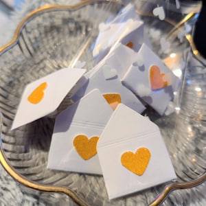Peut inclure: Une collection de petites enveloppes en papier blanc, chacune orn&eacute;e d'un c&oelig;ur en feutre dans des tons orange et p&ecirc;che. Les enveloppes sont dispos&eacute;es dans un sac en plastique transparent, dans un bol en verre d&eacute;coratif avec un bord dor&eacute;.