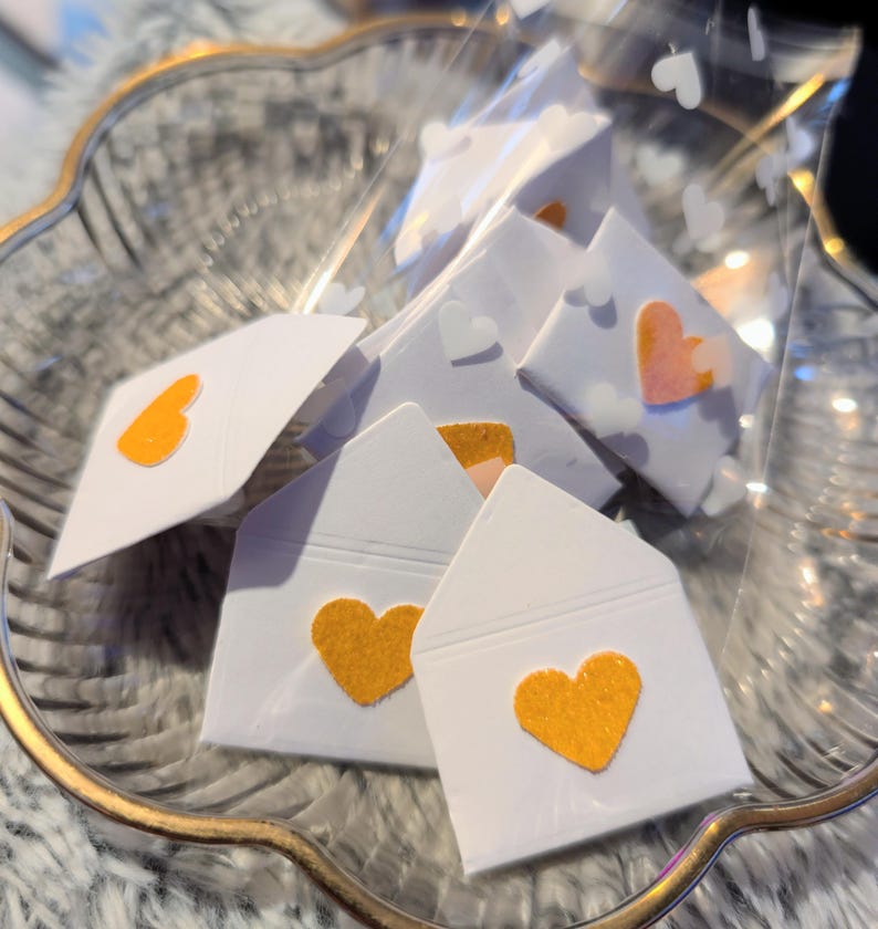 Peut inclure: Une collection de petites enveloppes en papier blanc, chacune orn&eacute;e d'un c&oelig;ur en feutre dans des tons orange et p&ecirc;che. Les enveloppes sont dispos&eacute;es dans un sac en plastique transparent, dans un bol en verre d&eacute;coratif avec un bord dor&eacute;.