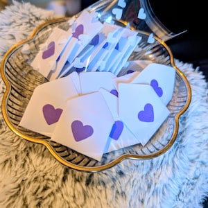Peut inclure: Un plat en verre transparent d&eacute;coratif avec un bord dor&eacute; contient plusieurs petites enveloppes blanches, chacune orn&eacute;e d'un c&oelig;ur violet. Certaines enveloppes sont dans un sac en plastique transparent avec des d&eacute;coupes en forme de c&oelig;ur. Le plat repose sur une surface blanche et duveteuse.