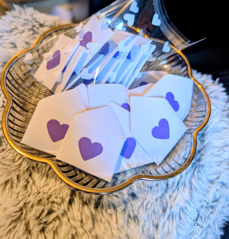 Peut inclure: Un plat en verre transparent d&eacute;coratif avec un bord dor&eacute; contient plusieurs petites enveloppes blanches, chacune orn&eacute;e d'un c&oelig;ur violet. Certaines enveloppes sont dans un sac en plastique transparent avec des d&eacute;coupes en forme de c&oelig;ur. Le plat repose sur une surface blanche et duveteuse.