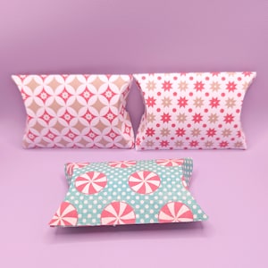 May include: Three rectangular pillow boxes with curved edges. The top left box features a pink and beige geometric floral pattern. The top right box has a pink and beige star and dot pattern. The bottom box displays a teal and pink peppermint candy print.