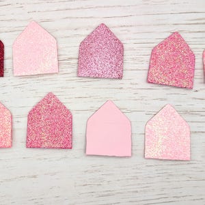 May include: A top-down view shows ten house-shaped decorations in shades of pink and red glitter. One is a solid deep red, others range from light pink to glittery rose. They are arranged on a light wood-grain surface.